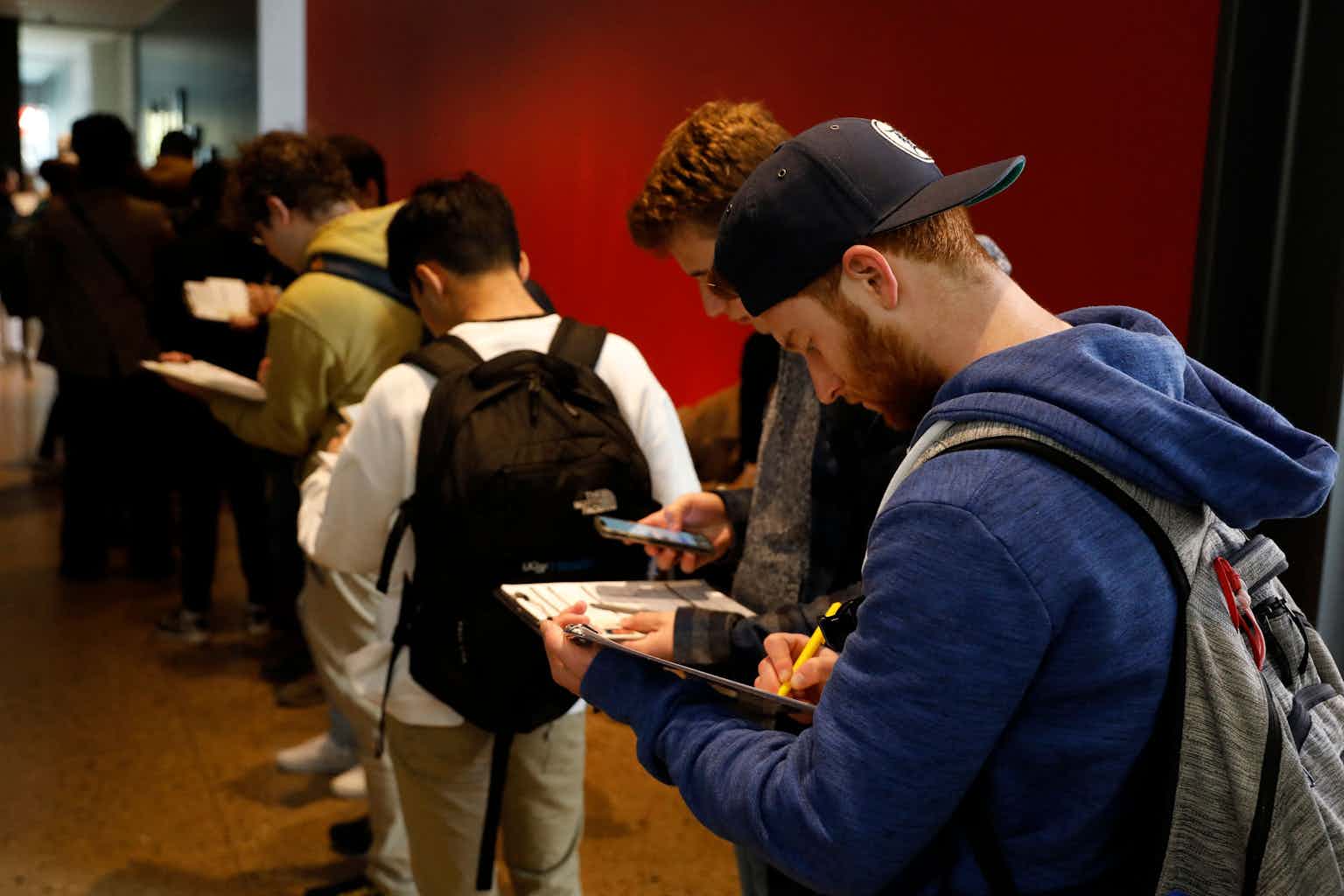 A row of young people stand inside a room with a red wall and look down at clipboards and white paper.