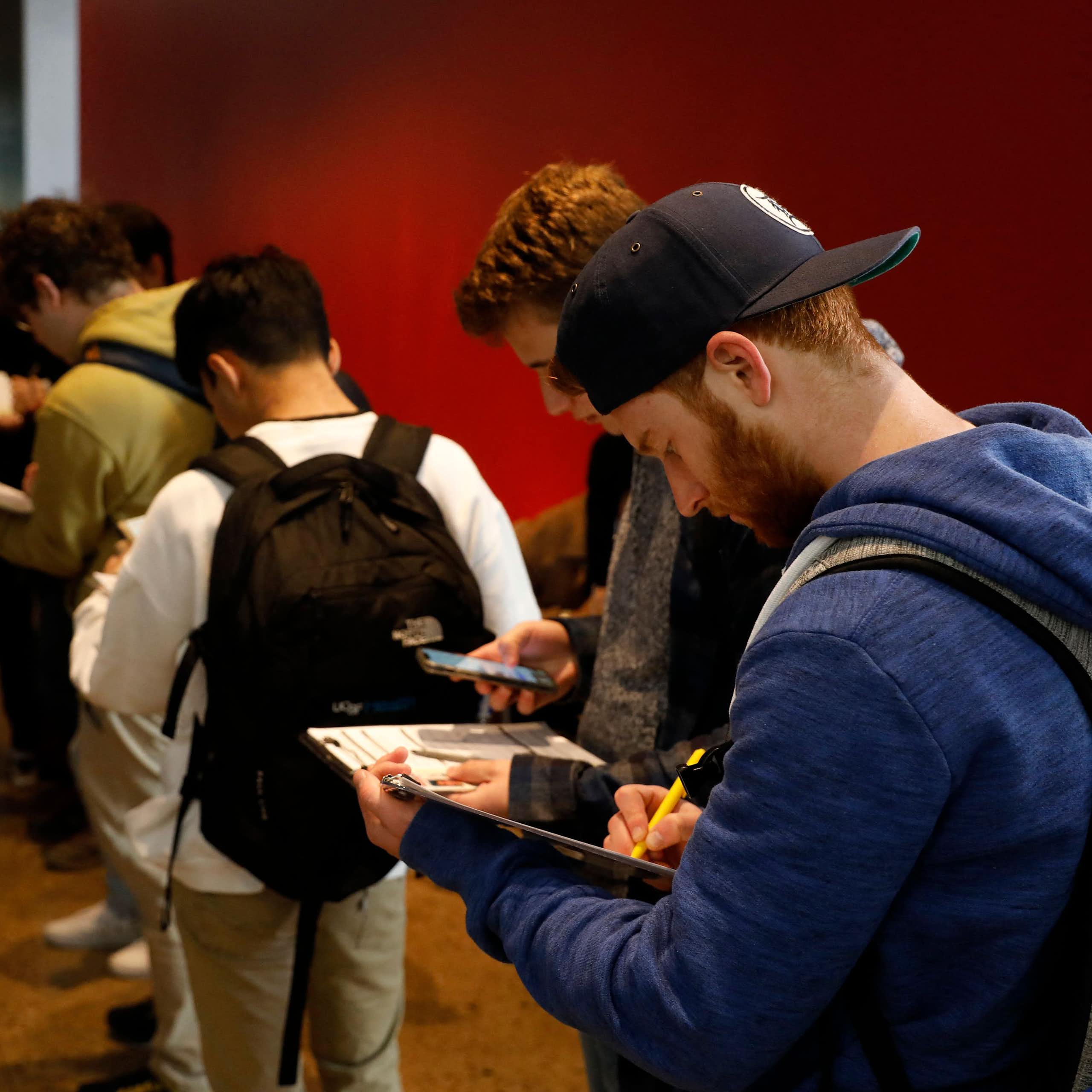 A row of young people stand inside a room with a red wall and look down at clipboards and white paper.
