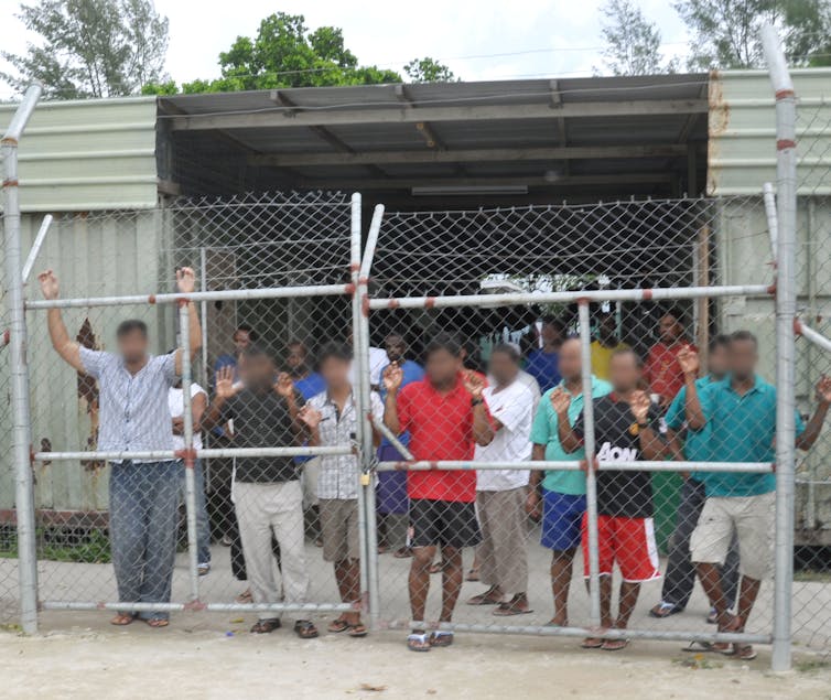 A row of men with blurred out faces stand behind a wire fence