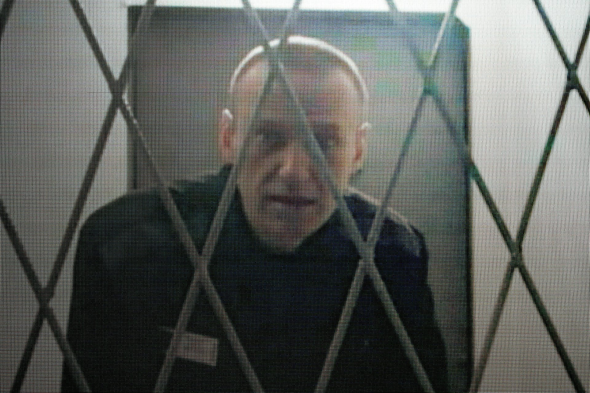 A thin man looks through prison bars.