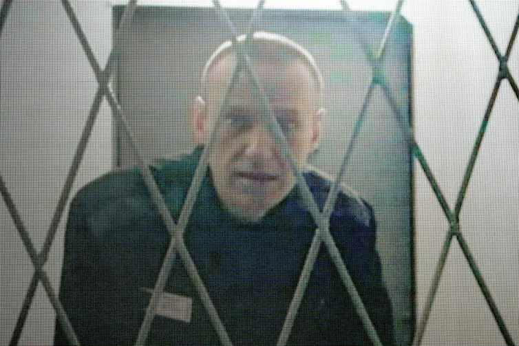 A thin man looks through prison bars.
