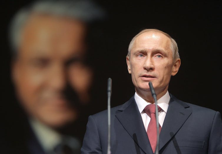 A balding man speaks into a microphone with the blurred image of an older man behind him on a screen.