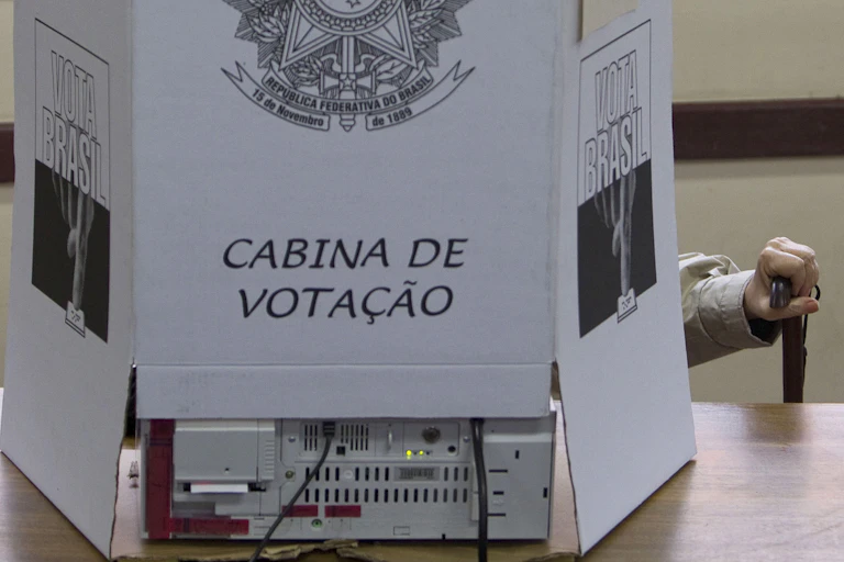Mão de um homem idoso segura uma bengala numa cabine de votação brasileira