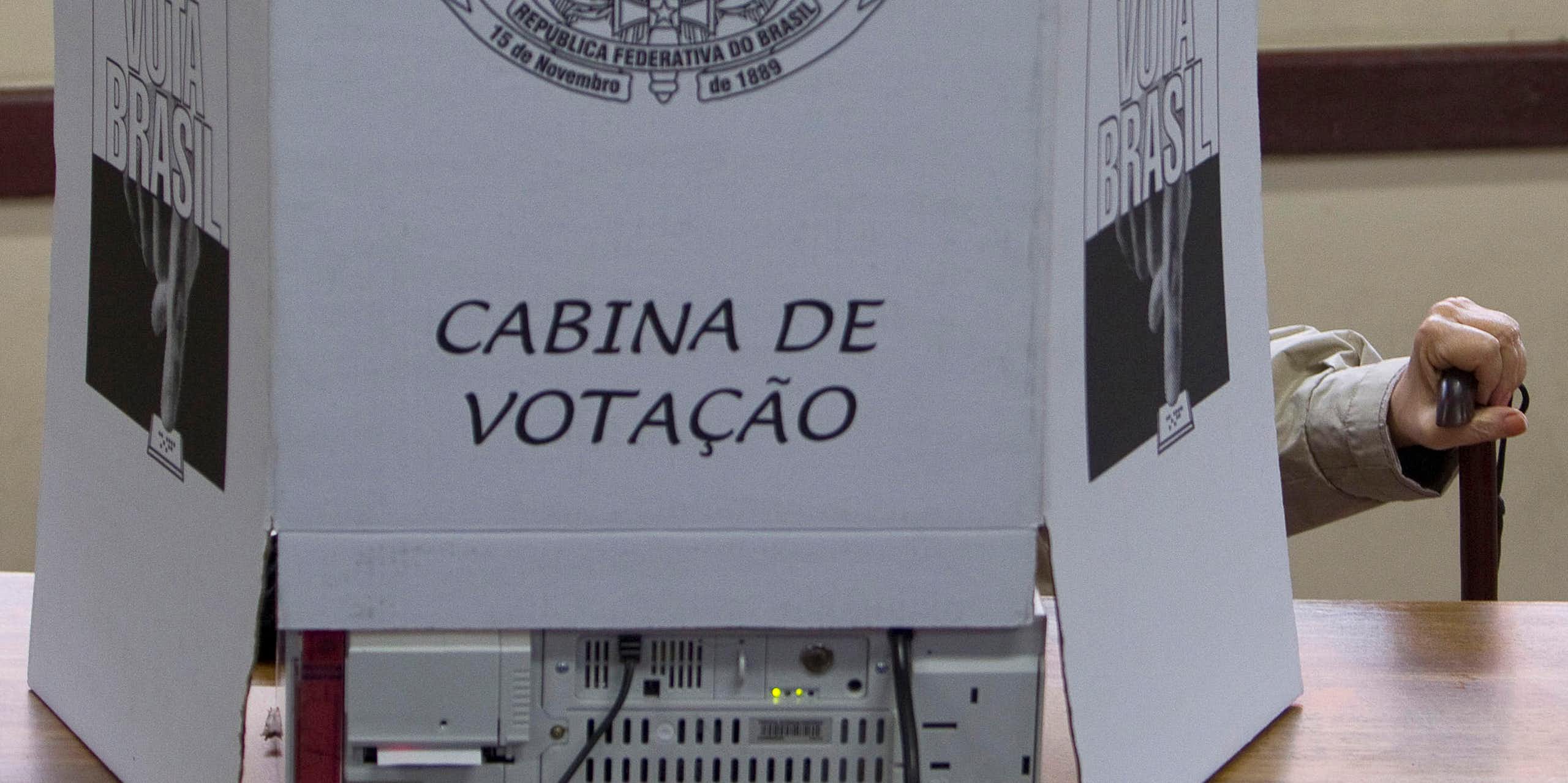 Mão de um homem idoso segura uma bengala numa cabine de votação brasileira