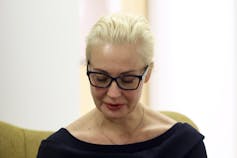 A blonde woman wearing glasses looks down.