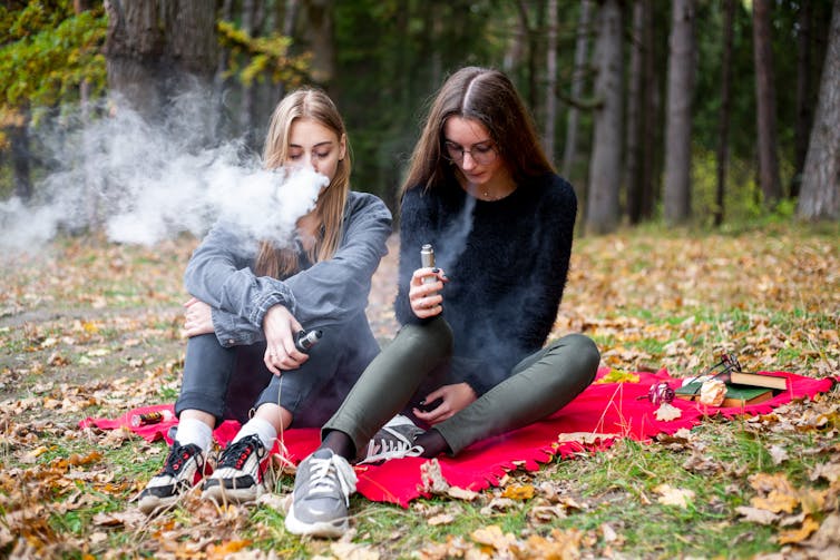 Two teenage girls vaping on a blanket in a park.