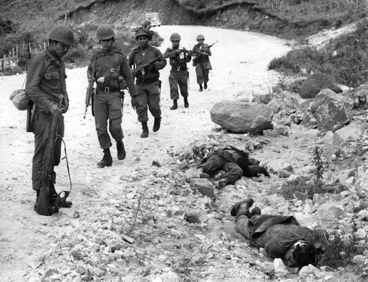 A black and white image of a group of soldiers looking at corpses next to a road.