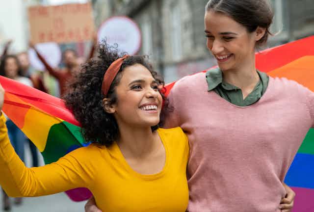 Two smiling young women with their arms around each other's waists carry a rainbow pride flag behind them
