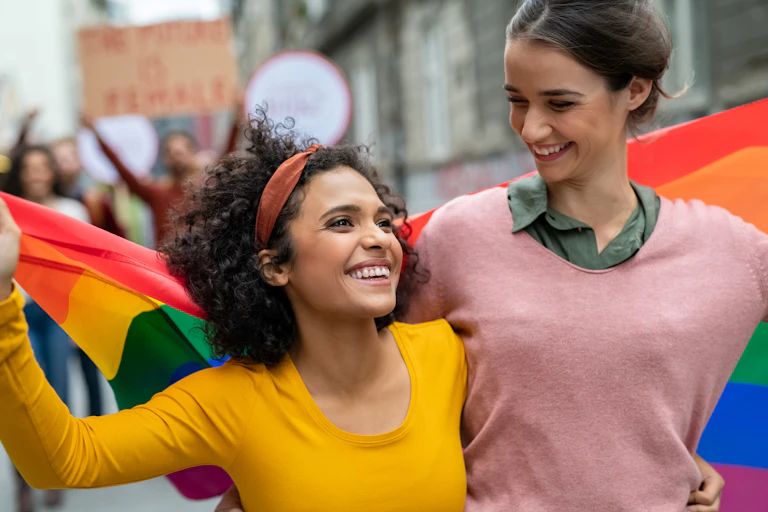 Two smiling young women with their arms around each other's waists carry a rainbow pride flag behind them