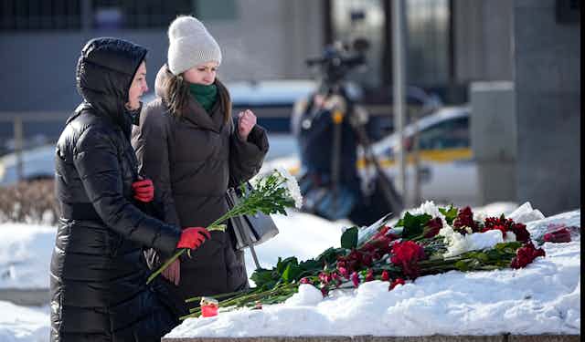Two women in long winter coats lay flowers on a snowy makeshift shrine at the foot of a monument.