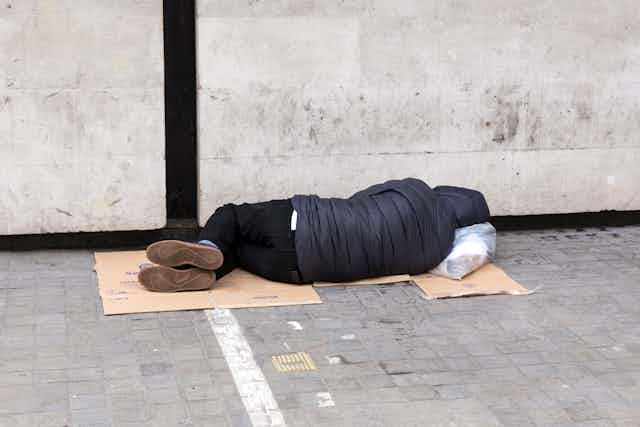 A homeless person sleeps on a piece of cardboard on the pavement.