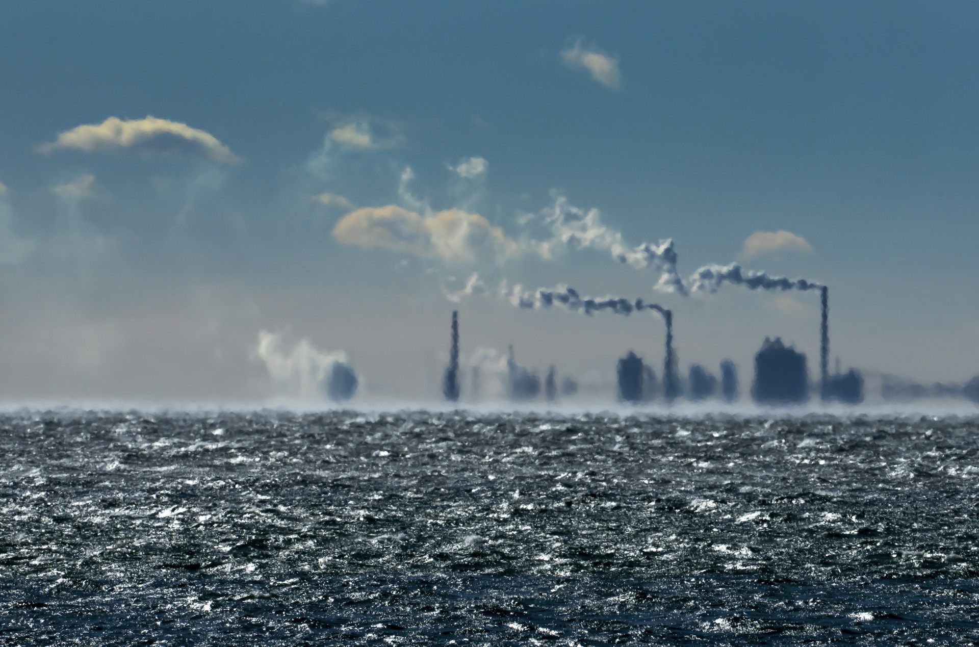 Hazy smoke stacks are seen in the distance with water in the foreground.