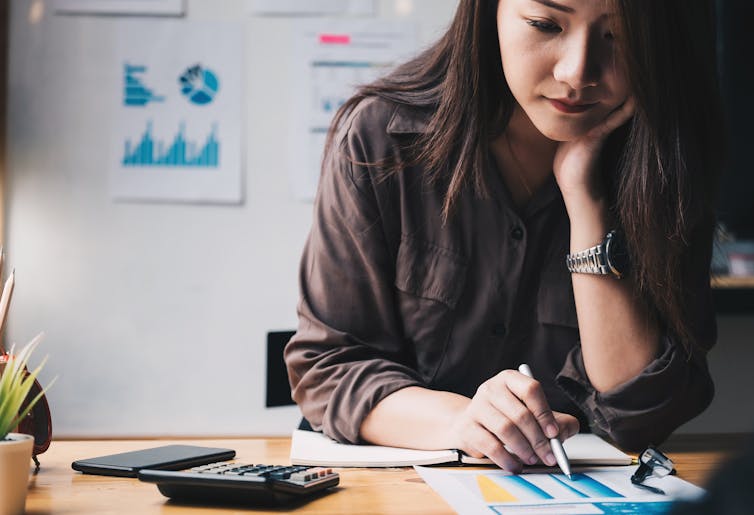 A young woman focuses, with her head resting on her chin at a desk, using a pen and calcuator to go through financial papers