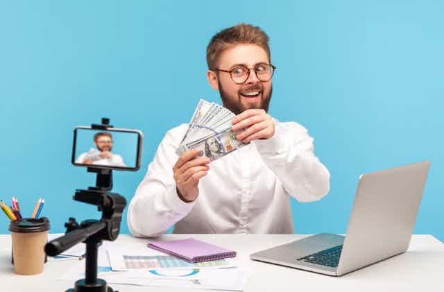 A young man in glasses and business shirt, sitting at a desk, films himself on a tripod, excitedly showing off a wad of cash
