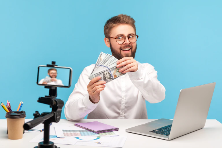 A young man in glasses and business shirt, sitting at a desk, films himself on a tripod, excitedly showing off a wad of cash