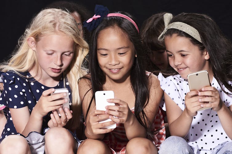 Three young girls sitting close together, each holding a smart phone.
