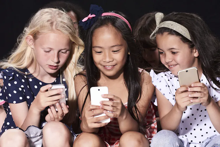 Three young girls sitting close together, each holding a smart phone.