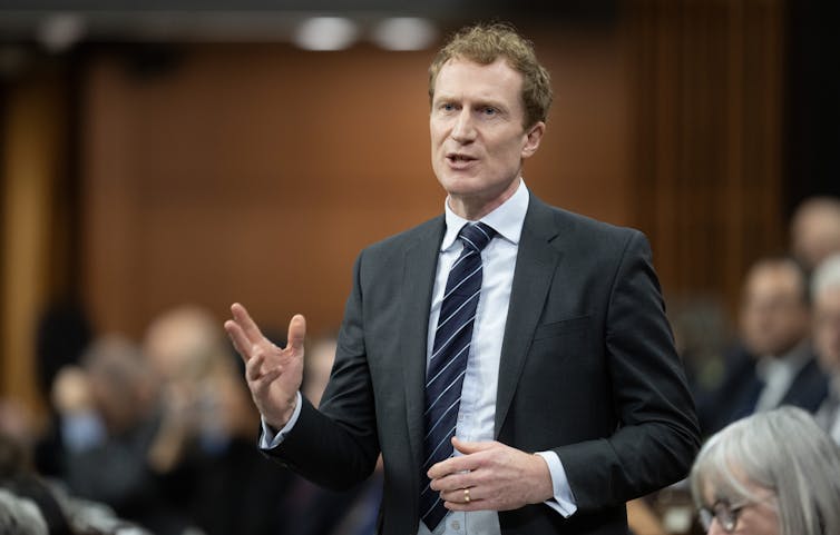 A middle-aged white man in a suit gestures with his hands while speaking to a room of people