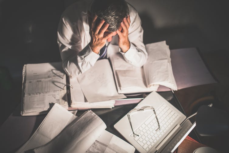 Person with head in hands, elbows planted on stacks of paperwork and books littering a desk, glasses and laptop on the side