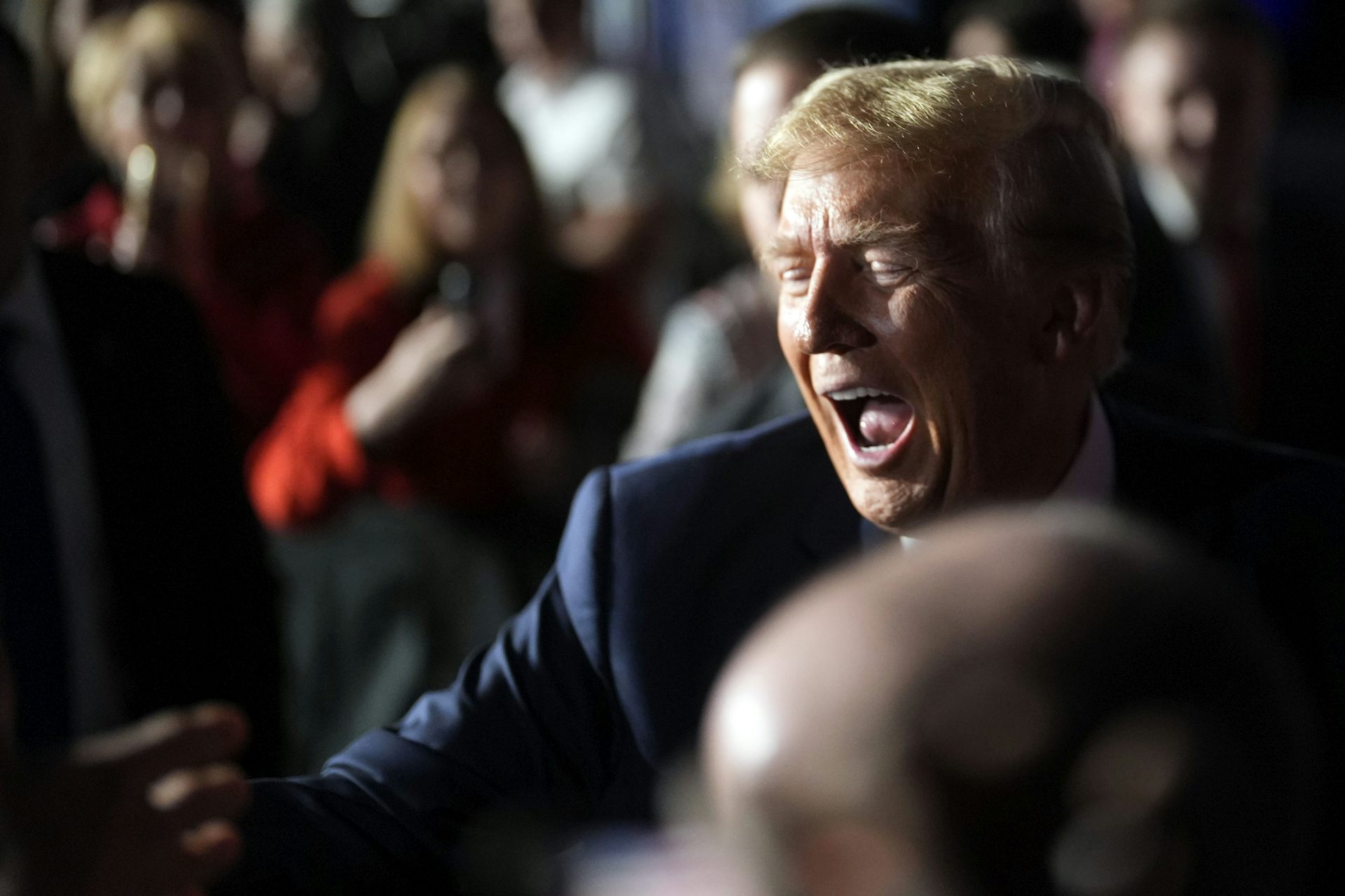 A man with golden hair makes a wide-mouthed face while speaking to people in a crowd.