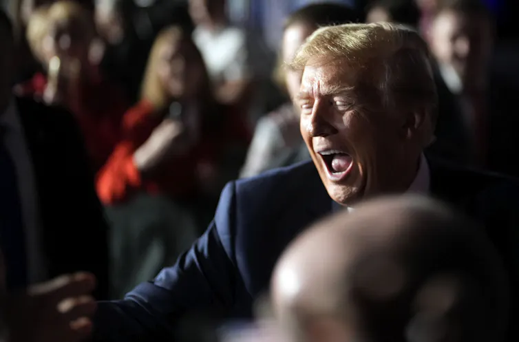 A man with golden hair makes a wide-mouthed face while speaking to people in a crowd.