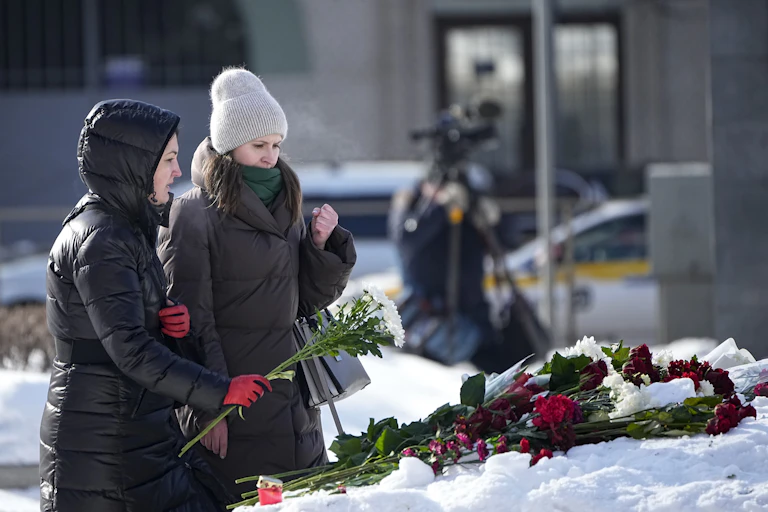 Deux femmes vêtues de longs manteaux d'hiver déposent des fleurs sur un autel de fortune enneigé, au pied d'un monument.