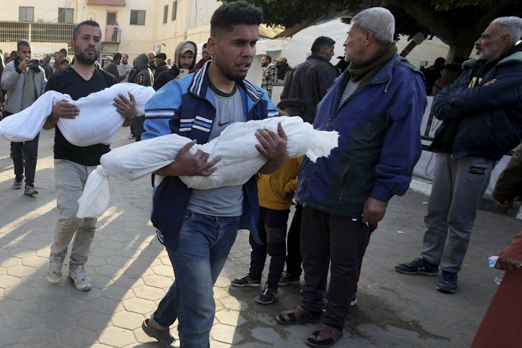 Two men carry the bodies of children shrouded in white cloth.