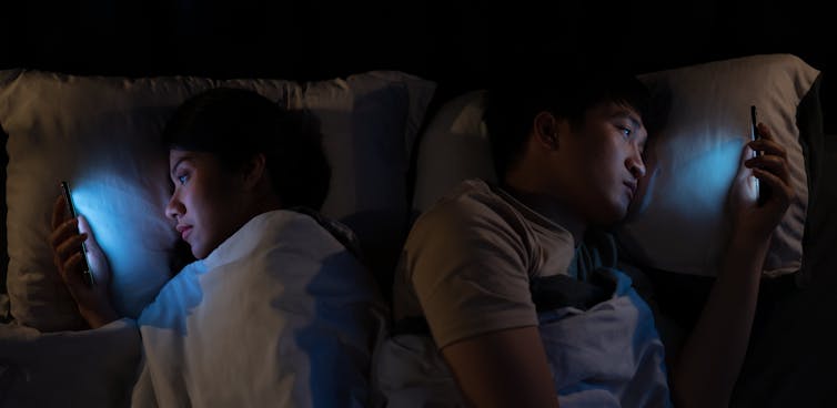Photo showing man and woman lying in bed in the dark, facing away from each other and looking at their phones.
