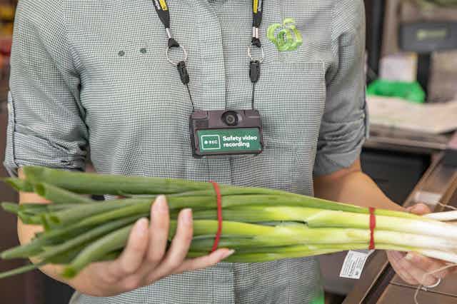 A photo of a Woolworths staff member wearing a camera labelled 'safety video recording' and holding a bunch of spring onions.