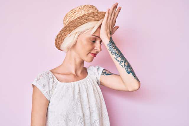 Young woman wearing hat, with tattooed arm, palm on forehead 