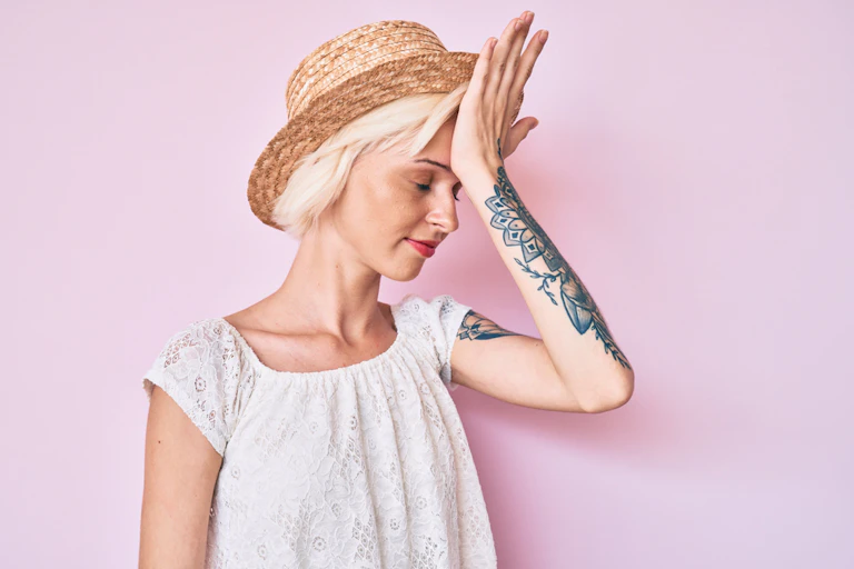 Young woman wearing hat, with tattooed arm, palm on forehead