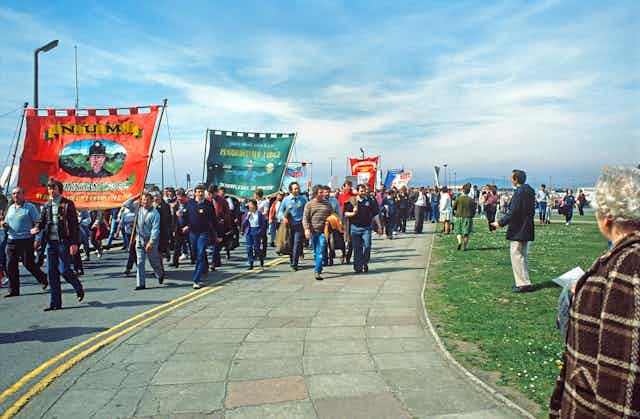 A large group of people holding huge banners march towards the camera on a fine day. 