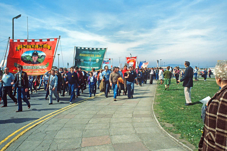 A large group of people holding huge banners march towards the camera on a fine day.