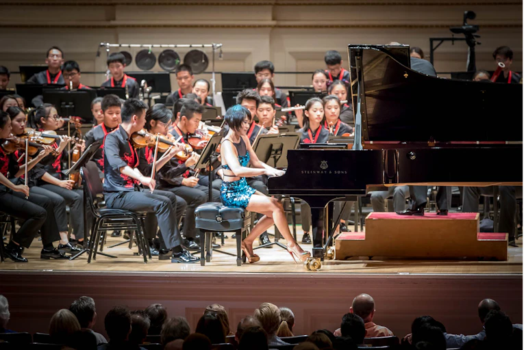 Una mujer joven toca el piano delante de una orquesta compuesta por chicos y chicas de unos veinte años.