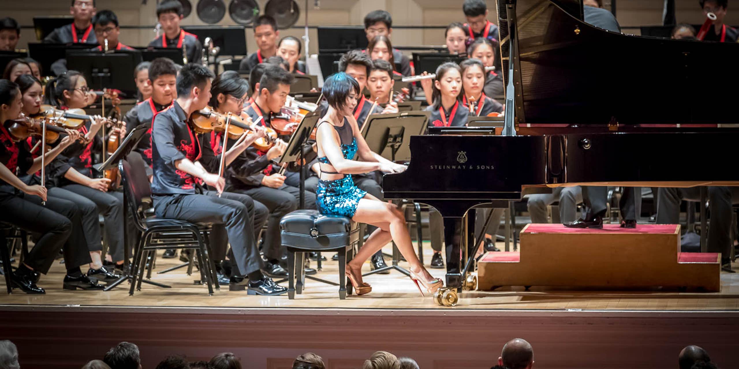 Una mujer joven toca el piano delante de una orquesta compuesta por chicos y chicas de unos veinte años.