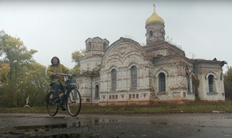 Woman rides a bike on a wet, cloudy day, past a damaged white church with gold dome.