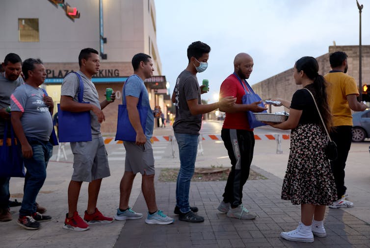 People, dressed mostly in shorts and T-shirts, stand in a line while a woman hands out packets to them.