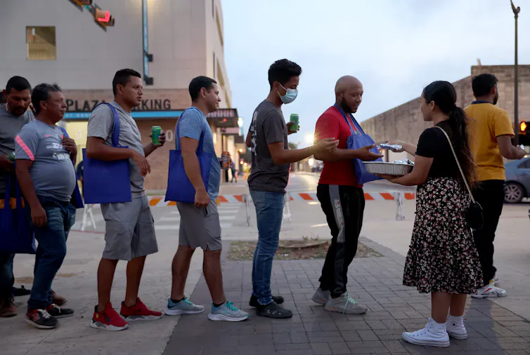 People, dressed mostly in shorts and T-shirts, stand in a line while a woman hands out packets to them.