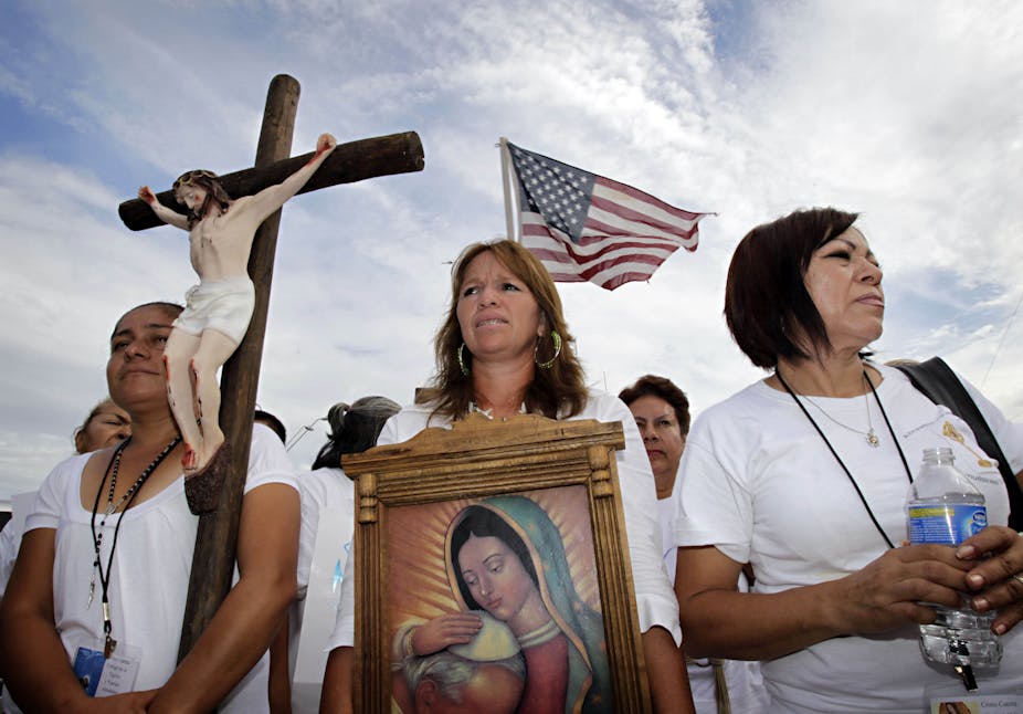 Three women stand ahead of a rally, with one holding a crucifix and another a portrait of Virgin Mary, while the American flag flutters in the back.