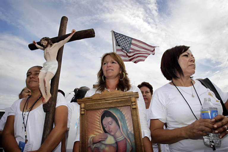 Three women stand ahead of a rally, with one holding a crucifix and another a portrait of Virgin Mary, while the American flag flutters in the back.