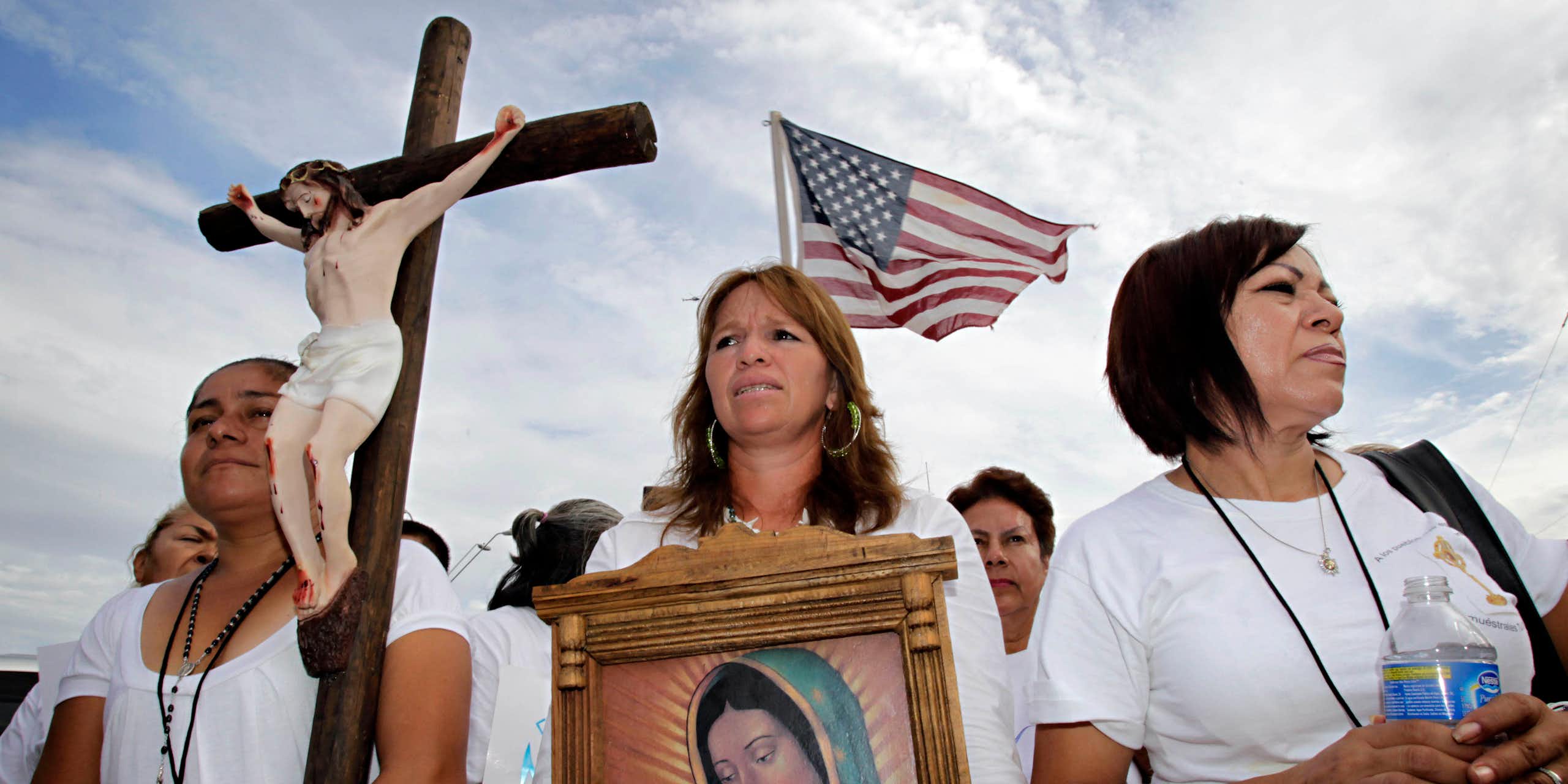Three women stand ahead of a rally, with one holding a crucifix and another a portrait of Virgin Mary, while the American flag flutters in the back.