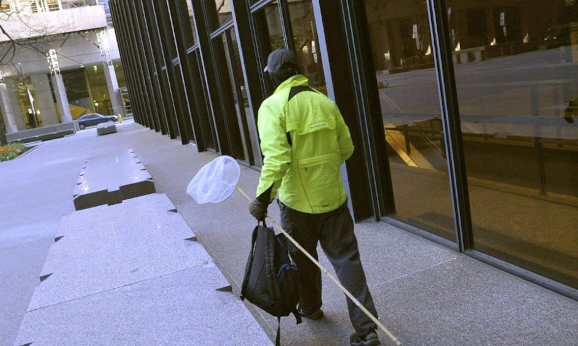 A person with a net walks in the shadow of a tall building next to some planters.
