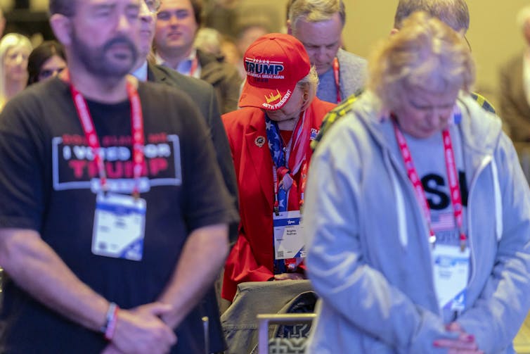 People wear red lanyards, shirts that say 'Trump' or 'USA' and hold their hands in front of them and bow their heads.