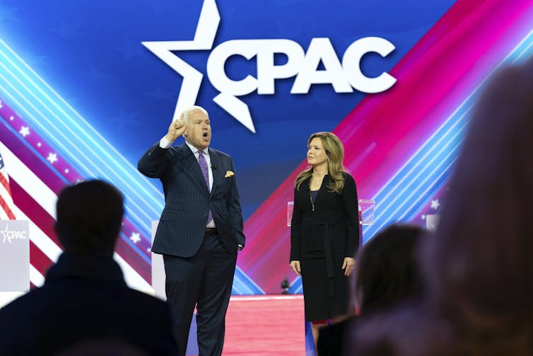 A white man with white hair and a dark suit stands on a stage with a woman in a black long sleeve dress. They stand in front of a large screen that is shades of red and blue and says 'CPAC' in white.