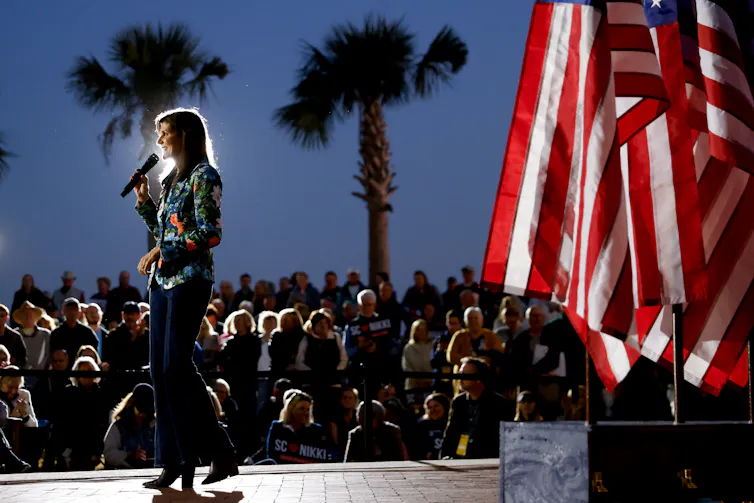 A tall, slim woman wears a long sleeve button down shirt and fancy pants and stands on a stage at twilight, with people in the crowd and palm trees in the distance. There are several American flags on the stage.