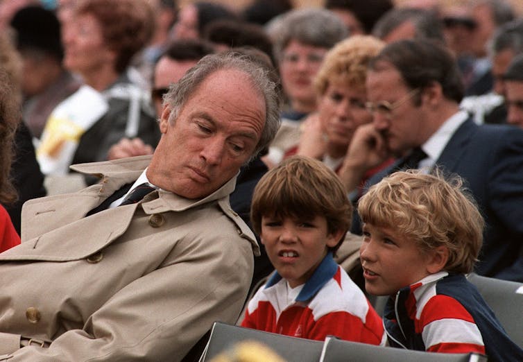 A man in a trench coat leans back and listens to one of two boys sitting behind him and talking to him during an event.
