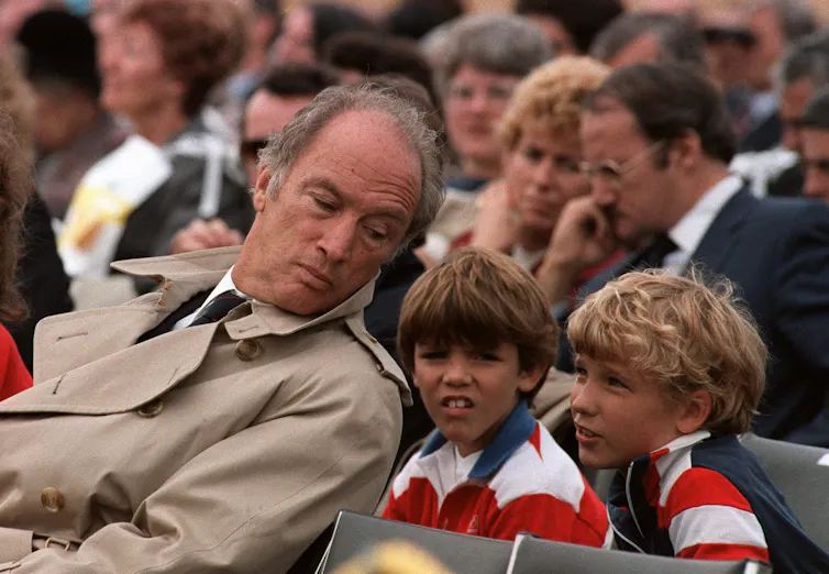 A man in a trench coat leans back and listens to one of two boys sitting behind him and talking to him during an event.