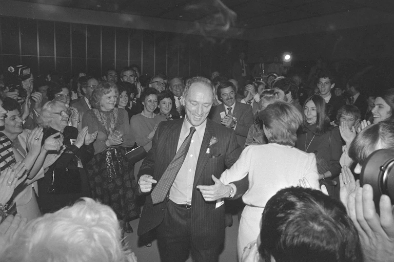 A man in a suit and tie smiles as he dances. People around him smile and dance.