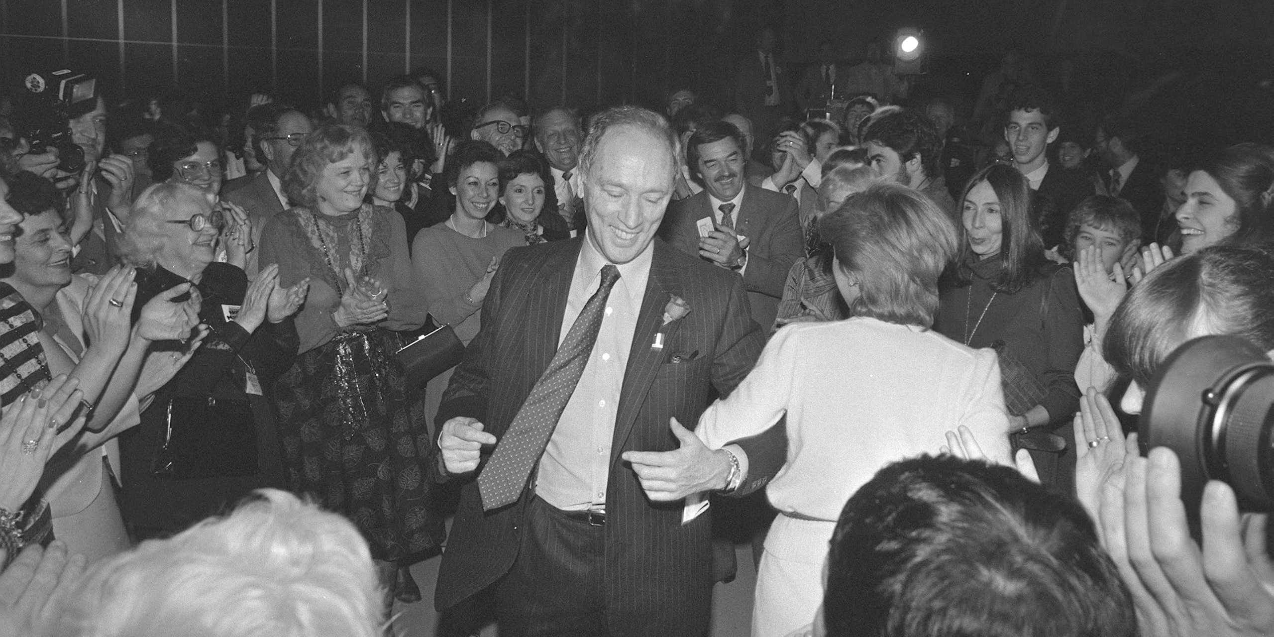 A man in a suit and tie smiles as he dances. People around him smile and dance.