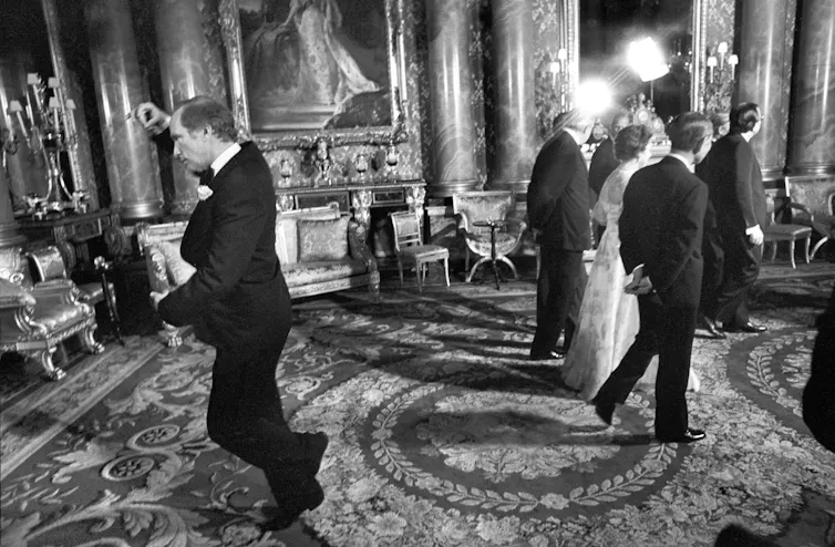 A black and white photo shows a man mid-pirouette as a woman in a formal ball gown has her back turned.